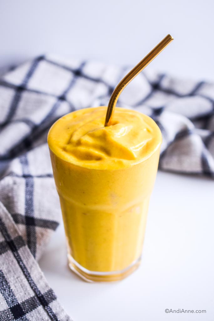 side view of mango turmeric smoothie in a glass with dark gold straw and striped kitchen towel behind it