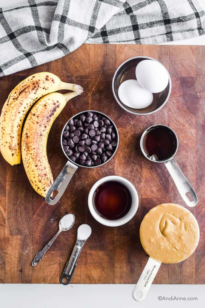 ingredients on wood cutting board. Includes ripe bananas, eggs, chocolate chips, maple syrup, vanilla, and seed butter