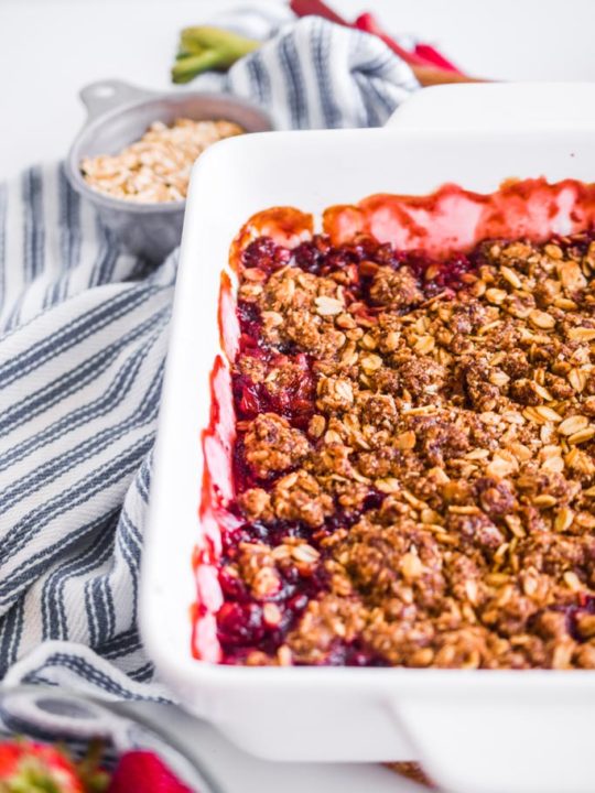 close up detail of strawberry rhubarb crisp in a white baking dish with striped kitchen towel beside it.