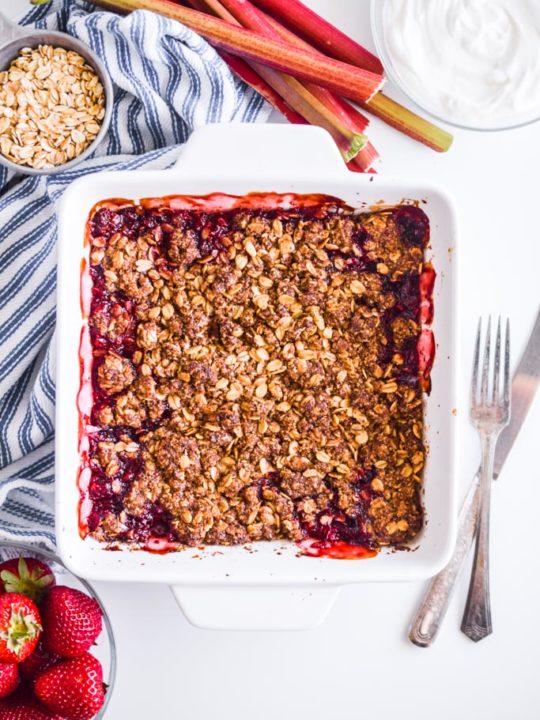 strawberry rhubarb crisp in a white baking dish with rhubarb, oats in a small bowl, yogurt in a small bowl, strawberries in a small bowl and fork and knife surrounding it