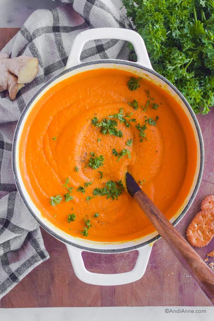turmeric ginger carrot soup in a white dutch oven with a wooden handle soup ladle. Parsley, ginger, crackers and a kitchen towel surround the soup pot.