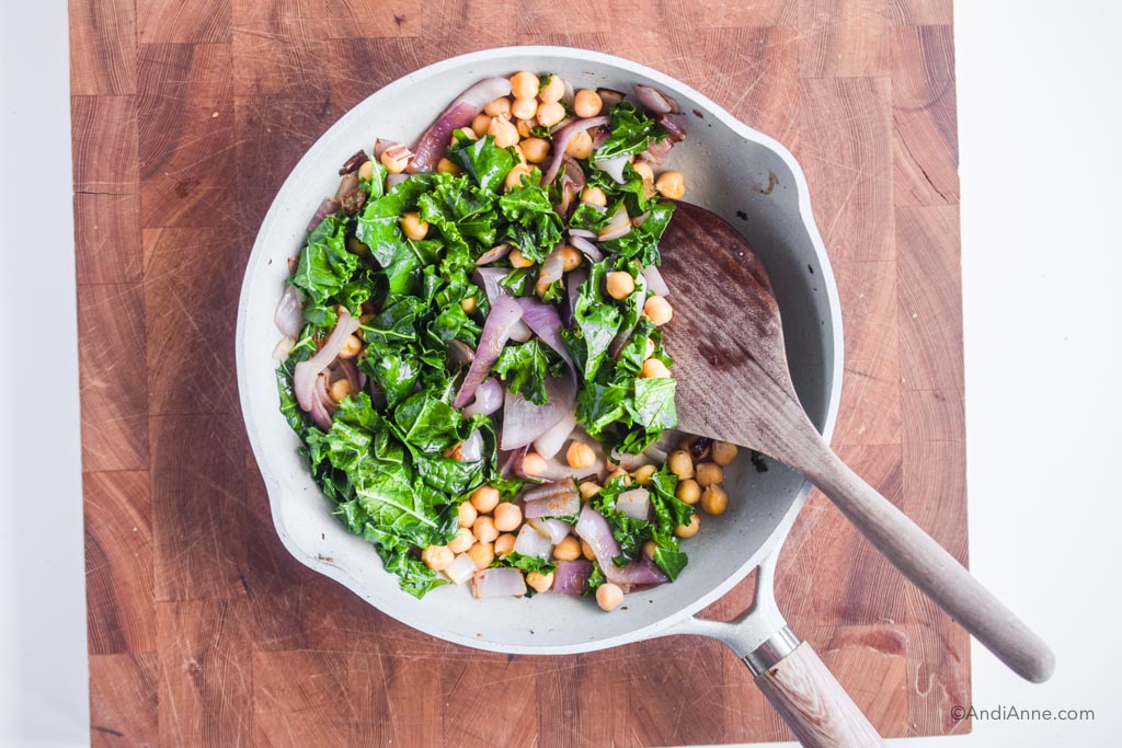 sauteed kale, chickpeas, and onions in a frying pan with a wooden spatula. Pan is sitting on a butcher block cutting board.