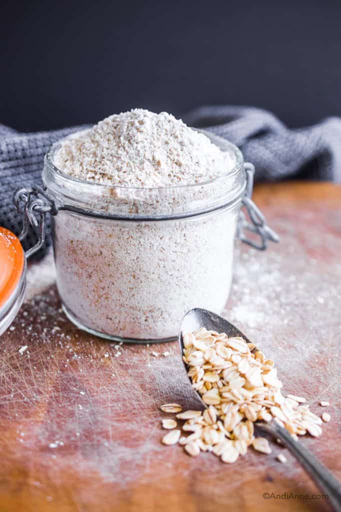 homemade oat flour in a glass jar. Rolled oats on a spoon in front and a dark kitchen towel in the background.