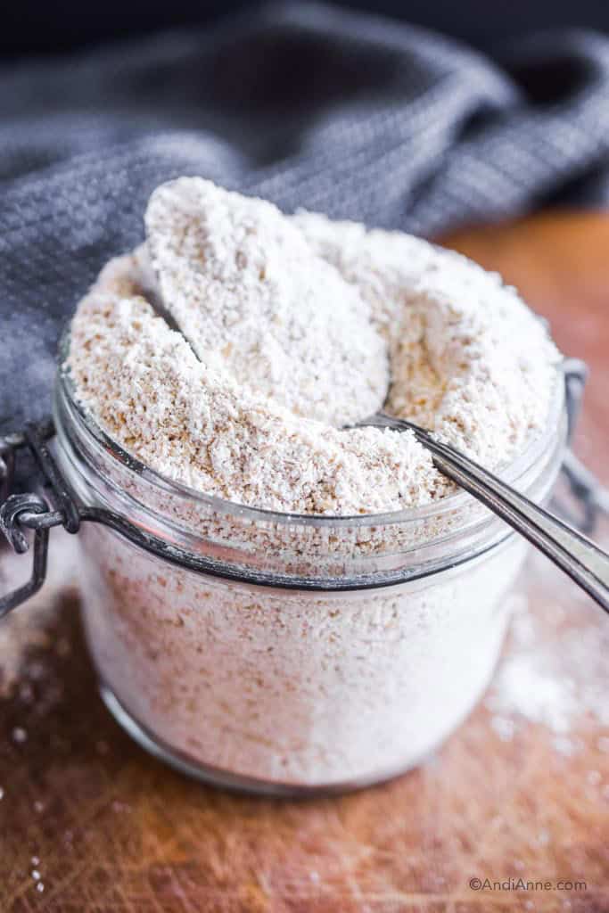 homemade oat flour in a glass jar with spoon on top. Dark kitchen towel in background.