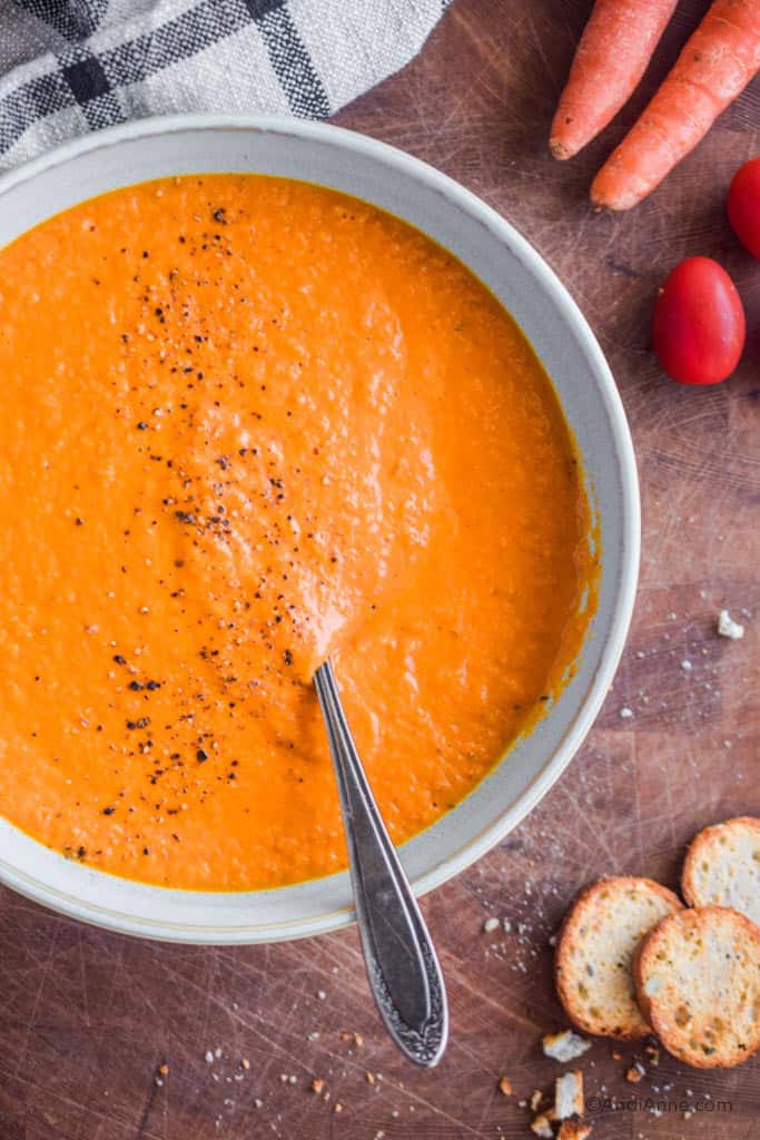 Pureed carrot tomato soup in bowl with spoon. Kitchen towel, carrots, tomatoes and crackers surround the bowl.