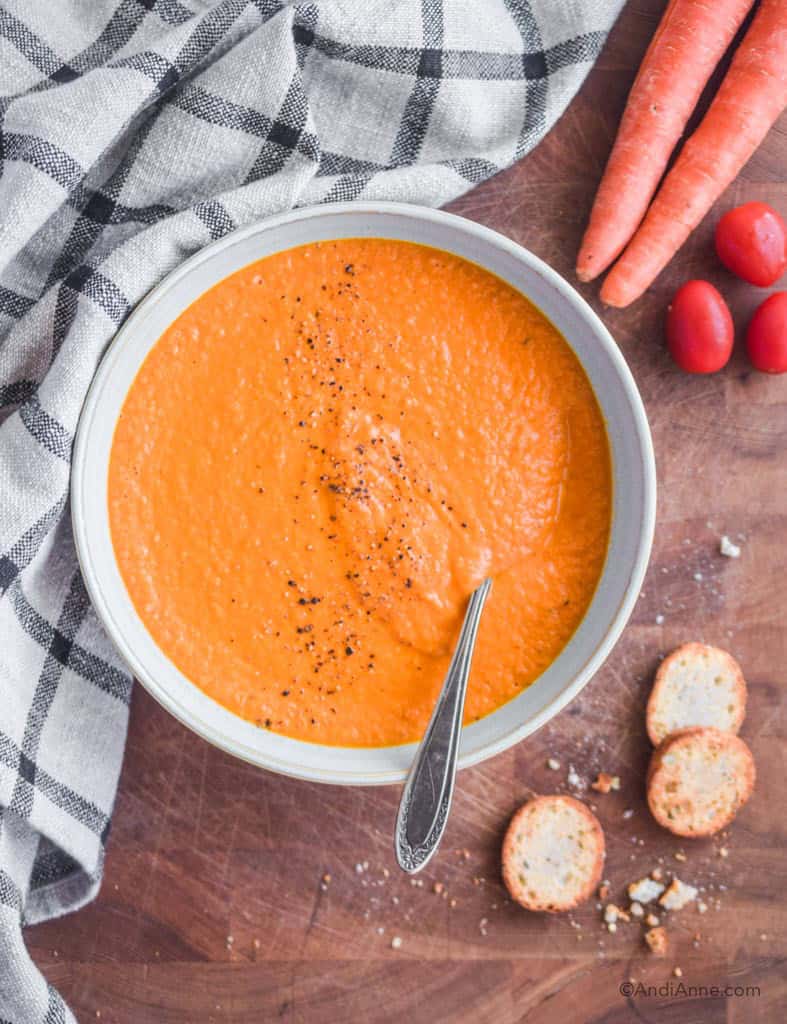 carrot tomato soup in bowl with spoon. A kitchen towel, carrots and tomatoes, and crumbled crackers surround the bowl.