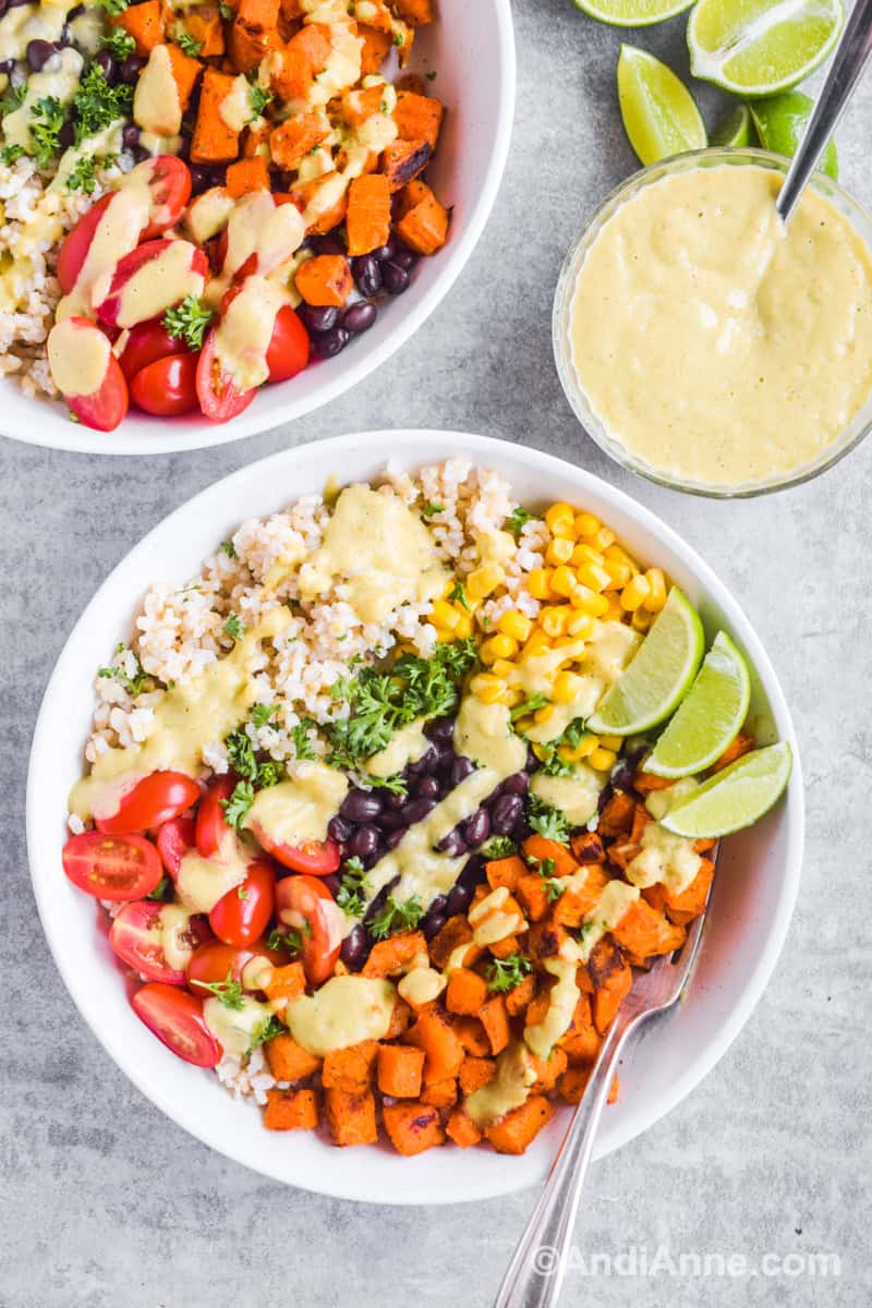 Two white bowls of baked sweet potatoe, rice, black beans, corn, cherry tomatoes, lime wedges and a fork. A bowl of green creamy dressing beside the plates.