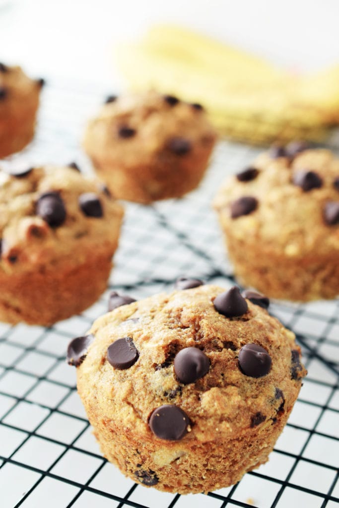 spelt muffin with banana and chocolate chips on a black cooling  rack. bananas blurred in background.