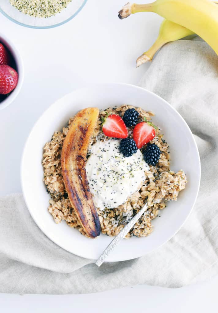 creamy oatmeal with fried banana in a white bowl with bananas, berries, and hemp seeds surrounding it