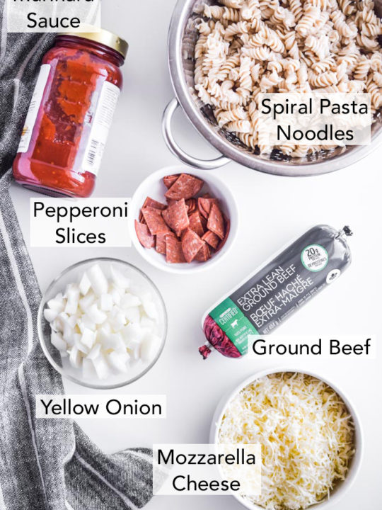 ingredients for the recipe in separate containers on a white table, including pepperoni slices in a white bowl, spiral pasta noodles, marinara, uncooked ground beef in a tube, chopped onion, and sliced mozzarella cheese