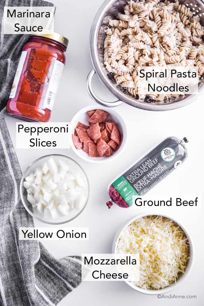 ingredients for the recipe in separate containers on a white table, including pepperoni slices in a white bowl, spiral pasta noodles, marinara, uncooked ground beef in a tube, chopped onion, and sliced mozzarella cheese