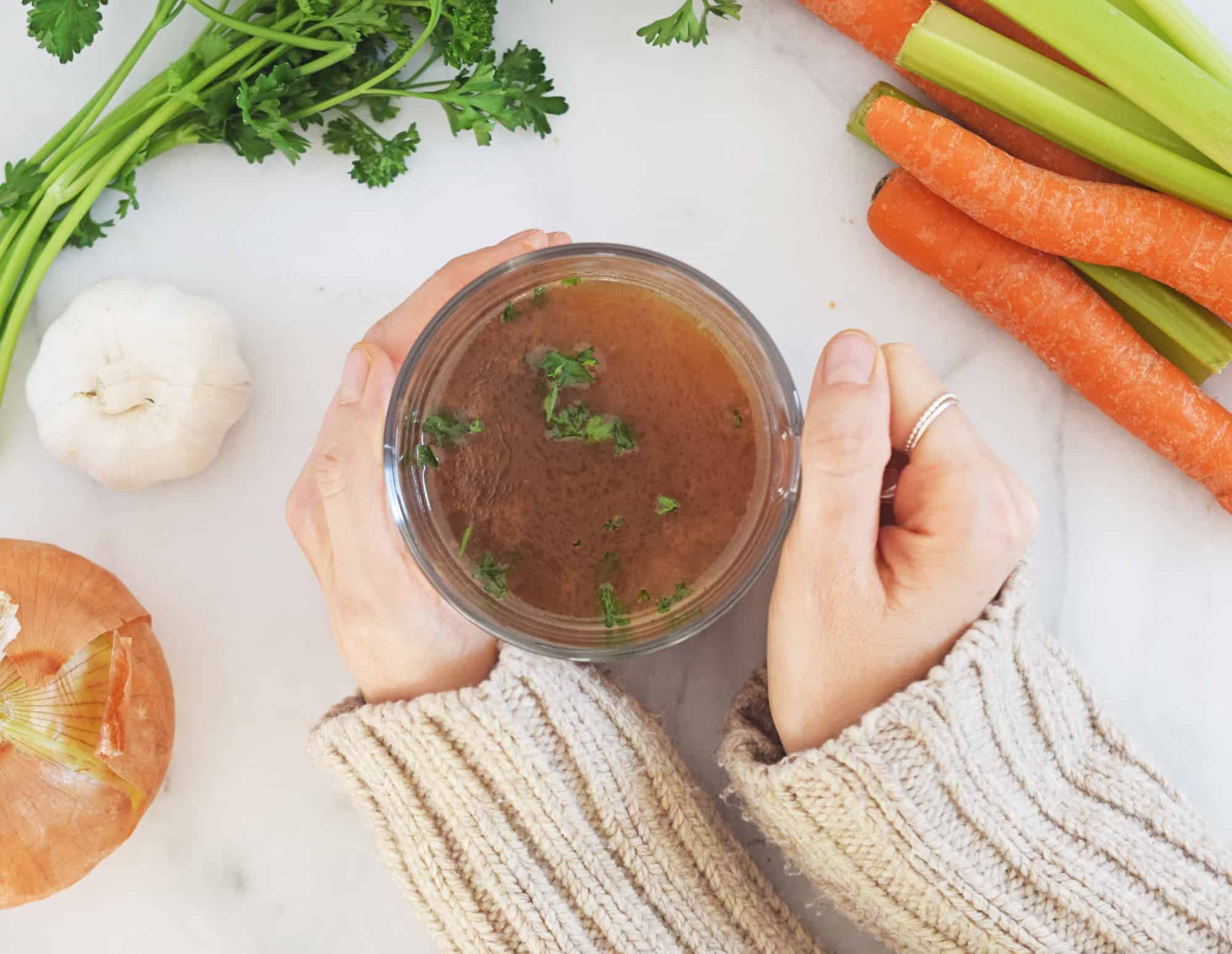 Hands holding a cup of homemade bone broth with parsley sprinkled on top. Carrots, celery, garlic and onion surround it.