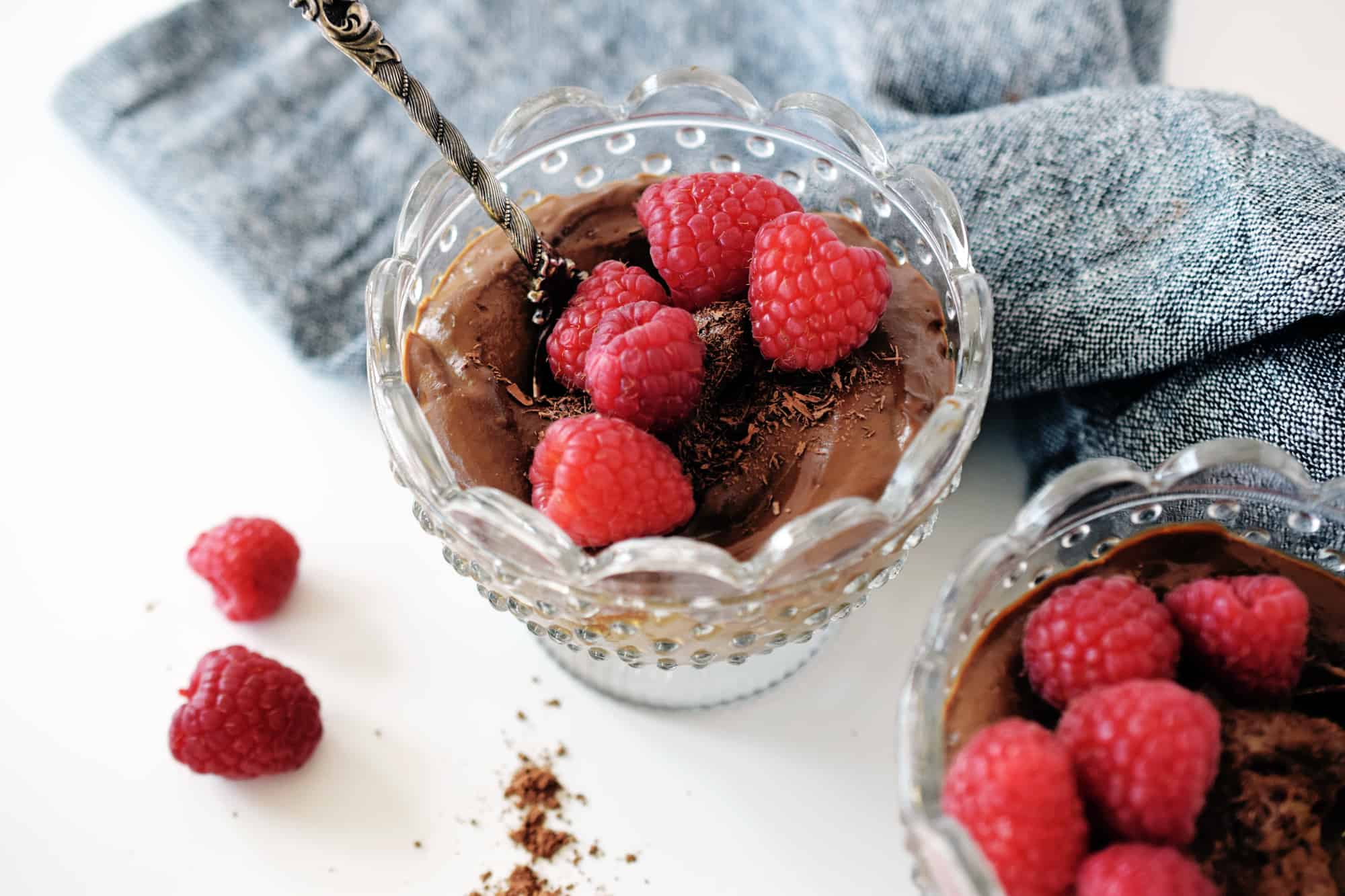 chocolate mousse in two glass bowls with fresh raspberries on top and spoon. Grey napkin in background