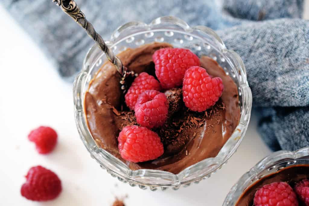 chocolate avocado pudding in a glass bowl with fresh raspberries on top and spoon. Grey napkin in background