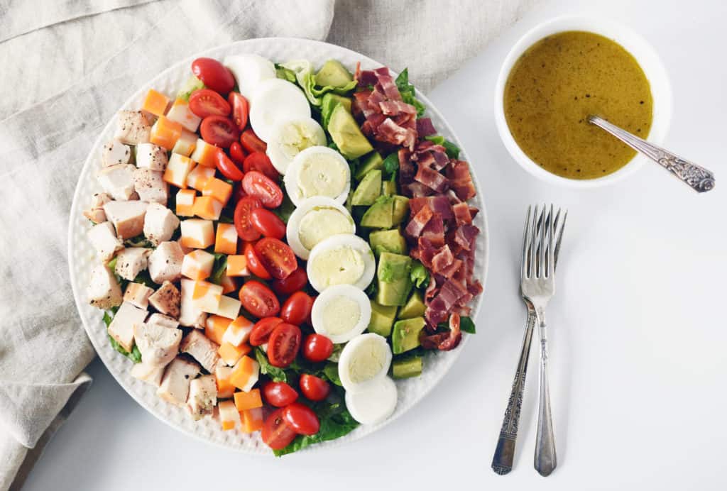cobb salad on a white plate, two forks bowl of salad dressing with spoon beside it