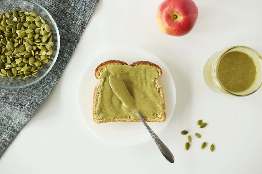 pumpkin seed butter spread on toast with knife. Apple and pumpkin seeds surround it on white counter.