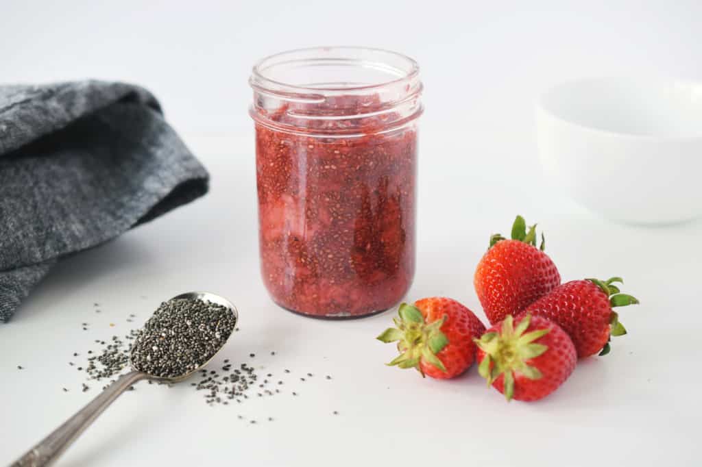 strawberry chia jam in mason jar. Spoon with chia seeds and four fresh strawberries are in foreground. Grey napkin and white bowl are beside mason jar.