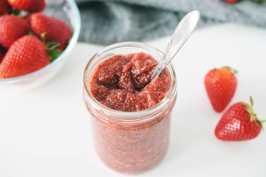 strawberry chia jam in mason jar with spoon. fresh strawberries in a bowl and dark grey napkin are in background.