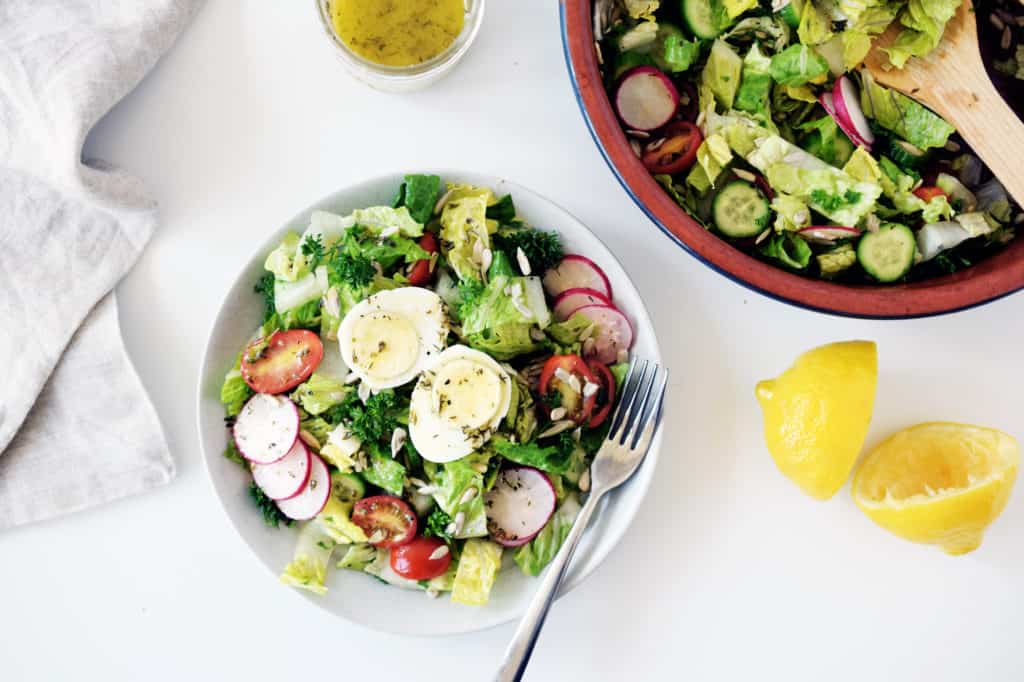 garden salad on a white plate with fork, lemon peels on the side and bowl of garden salad in top right