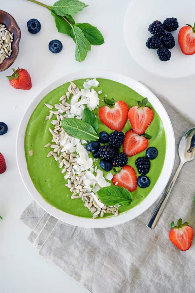 tropical green smoothie bowl with blueberries, strawberries, shredded coconut and sunflower seeds on top. Silver spoon on side with fresh berries sprinkled around.
