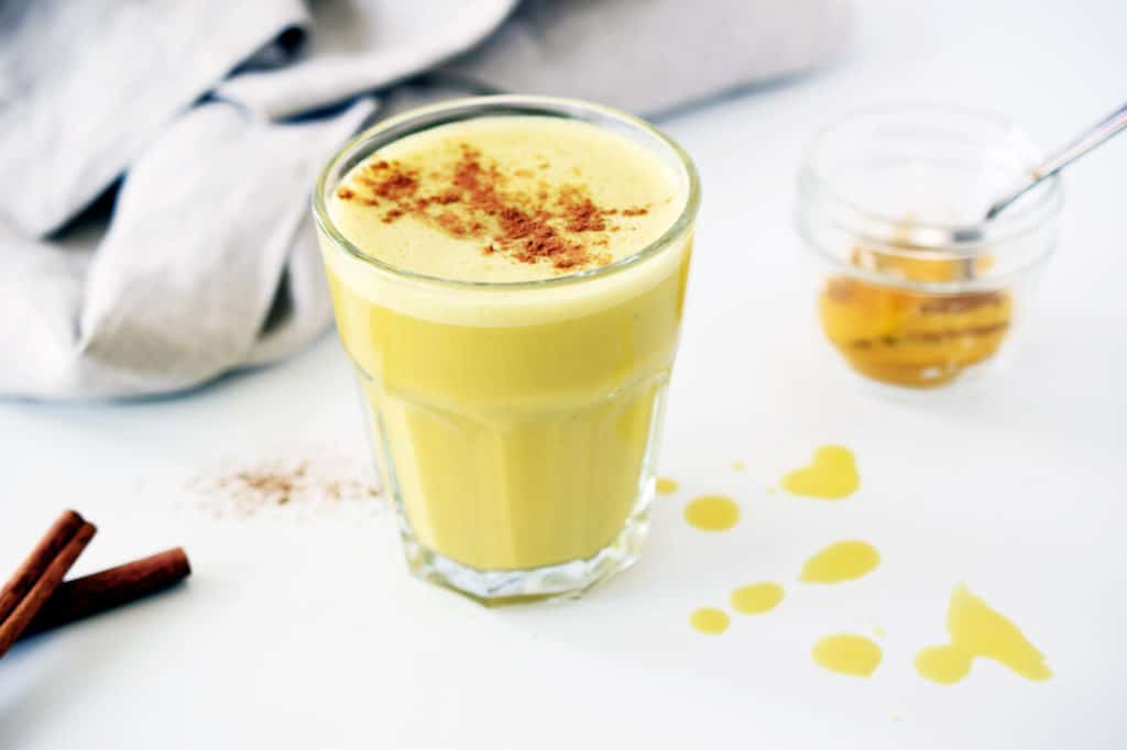 turmeric ginger latte in glass with napkin, cinnamon sticks and small mason jar with turmeric powder in background