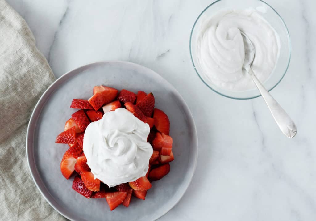 grey plate with sliced strawberries and whipped coconut cream on top. Glass bowl to the right with more cream inside and a spoon
