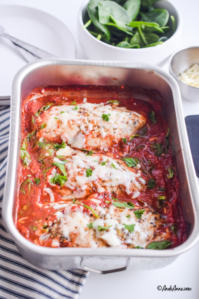 angled detail of stainless steel pan with mozzarella chicken recipe inside. White bowl with spinach and white plates are in background.