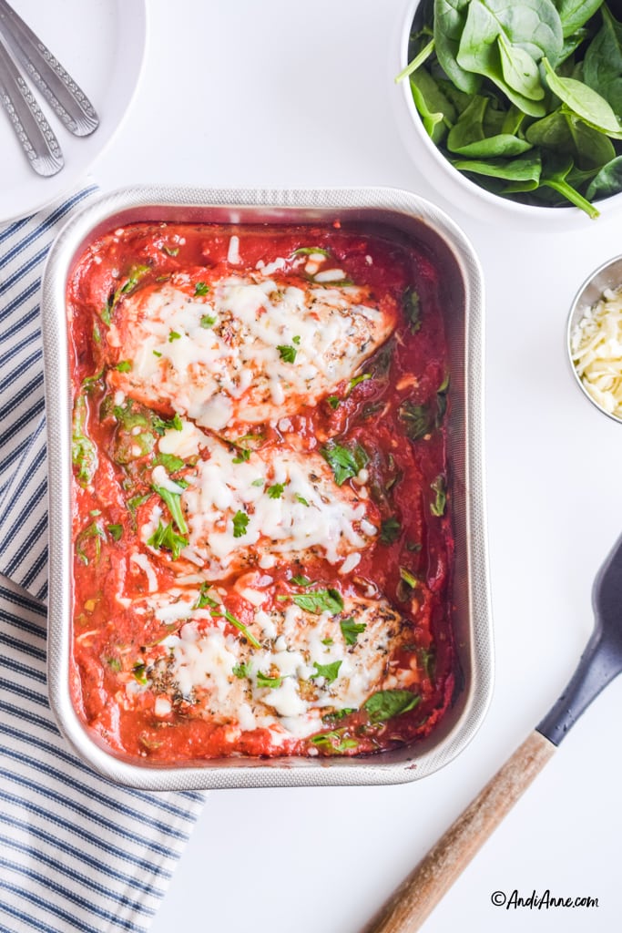 mozzarella chicken in tomato sauce in steel baking dish. A bowl of spinach, bowl of shredded mozzarella, as spatula and a striped napkin are surrounding the main dish.