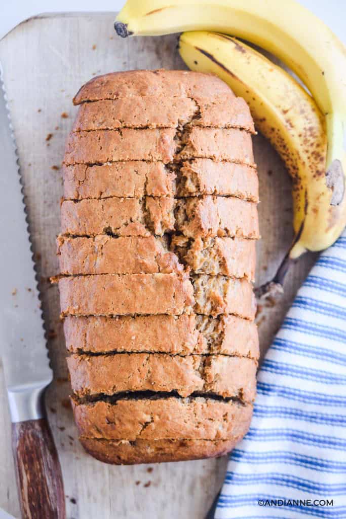 banana bread in thin slices on cutting board with bread knife and ripe bananas