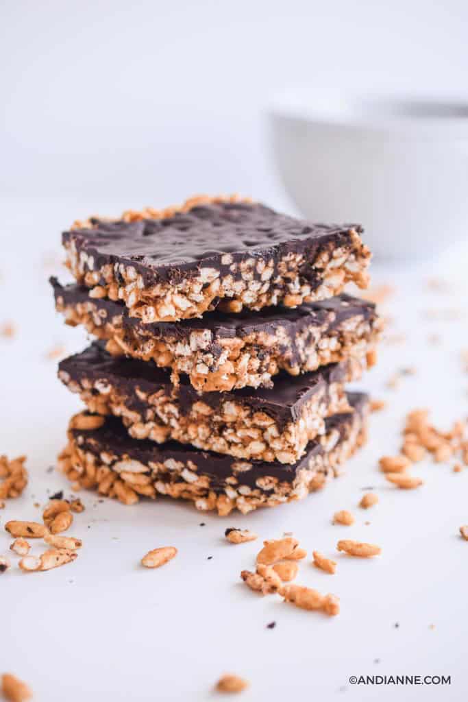 stacked chocolate puffed rice squares on top of eachother. White bowl in background. Puffed rice crumbles in foreground.