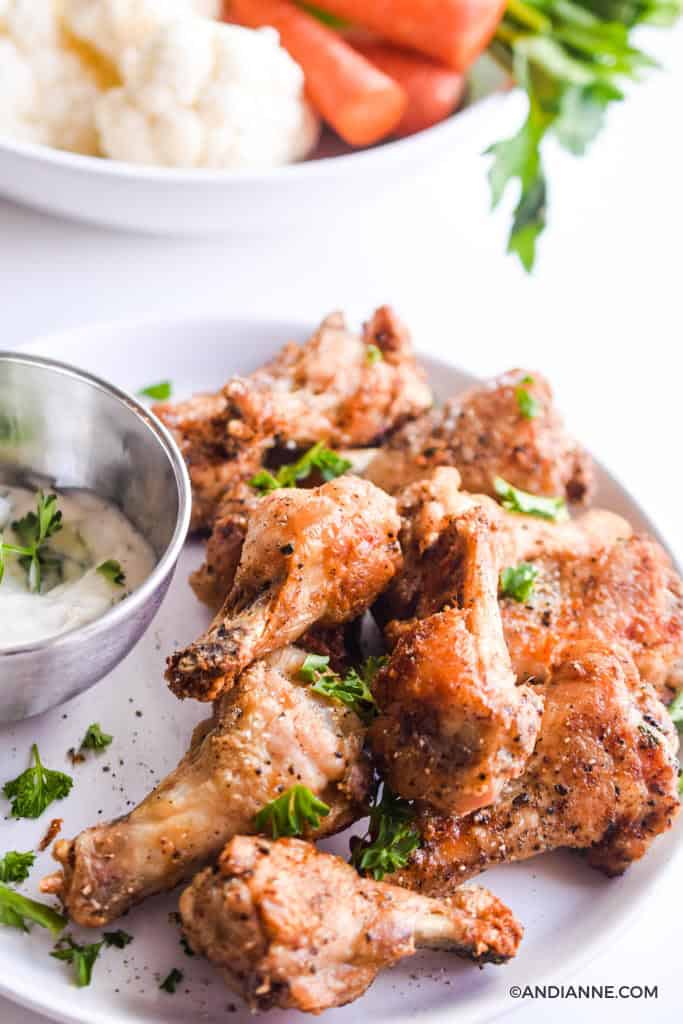 air fryer chicken drumettes on white plate with silver bowl of ranch dip on the left side. Cauliflower, sliced carrots, and sliced celery in a white bowl in the background.