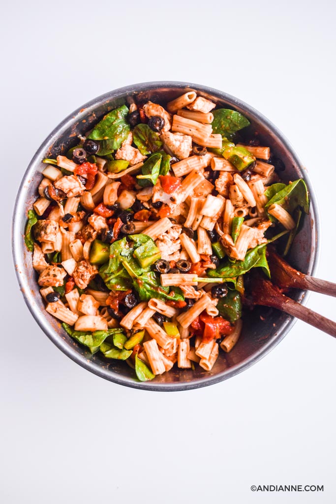 Cooked pasta, spinach, olives and tomatoes in a big steel bowl with two wooden spoons.