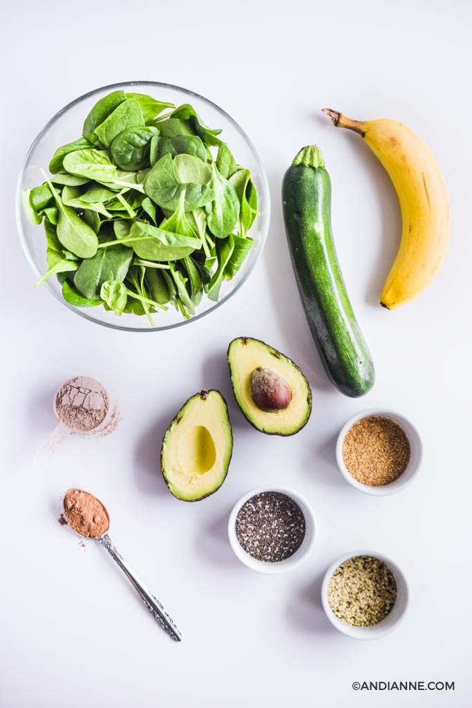 vegetables (spinach, zucchini and an avocado sliced in half), protein powder in spoons and seeds on a white counter in small bowls.