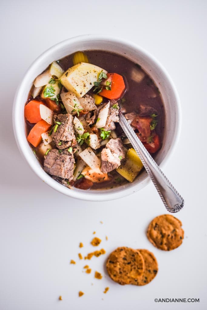 white bowl of beef stew with silver spoon. Three crackers on white counter with crumbs.