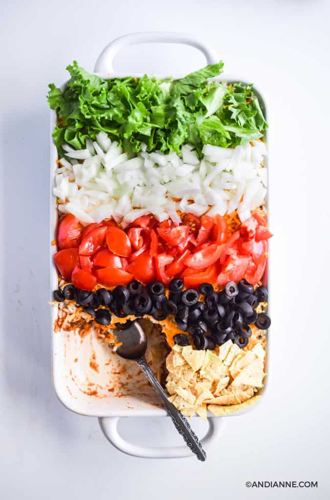 taco salad casserole with stripes of fresh vegetables and crushed nacho chips on top to create a rainbow effect. Once serving has been taken out of the bottom left corner. A silver spoon is in its place.