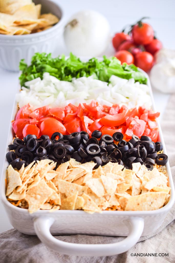 angled close up of taco casserole recipe in white dish with stripes of crushed nachos, sliced olives, sliced tomatoes, sliced onion, and chopped lettuce are on top.