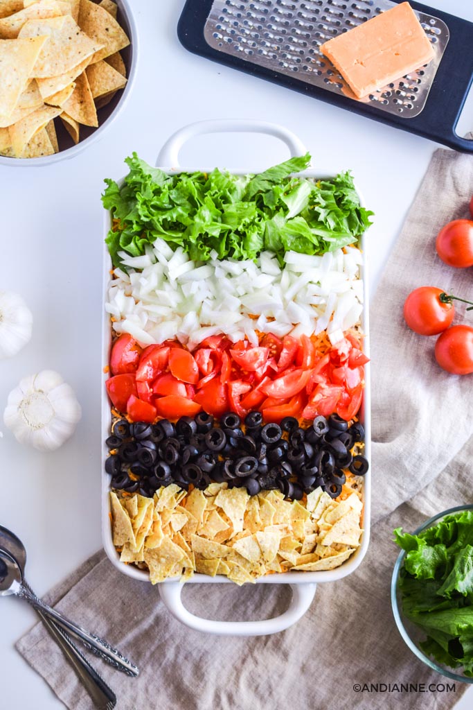 taco salad casserole with stripes of fresh vegetables and crushed nacho chips on top to create a rainbow effect. A cheese grater, bowl of nacho chips, fresh tomatoes, garlic bulbs and spoons all surround the casserole dish.