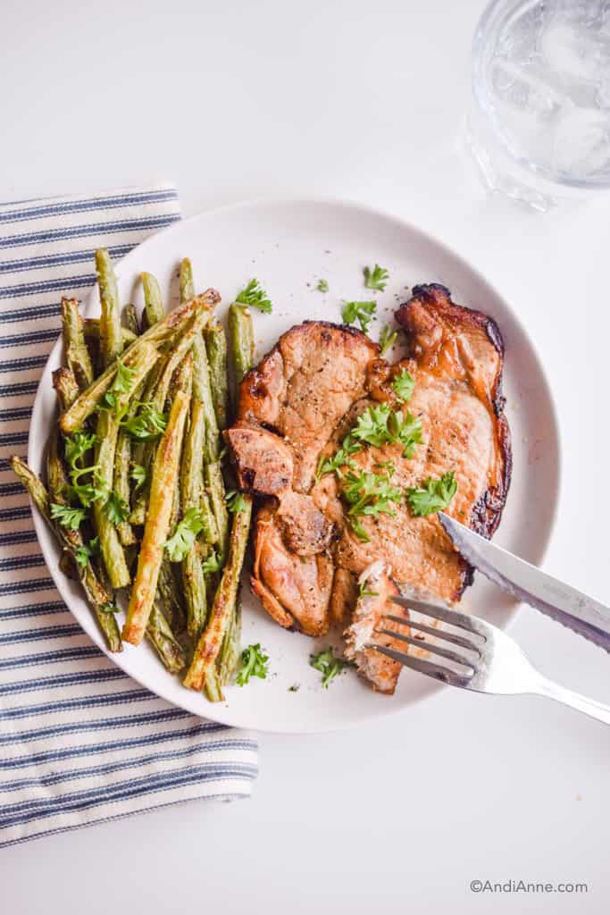 air fryer green beans and air fryer pork chop on white plate with fork and steak knife. Striped white and blue napkin on left.