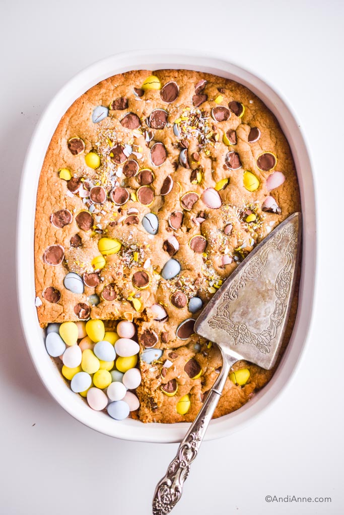 easter egg blondies cooked in white oval dish with silver pie cutter on top and cadbury mini eggs poured into cut out piece in bottom left