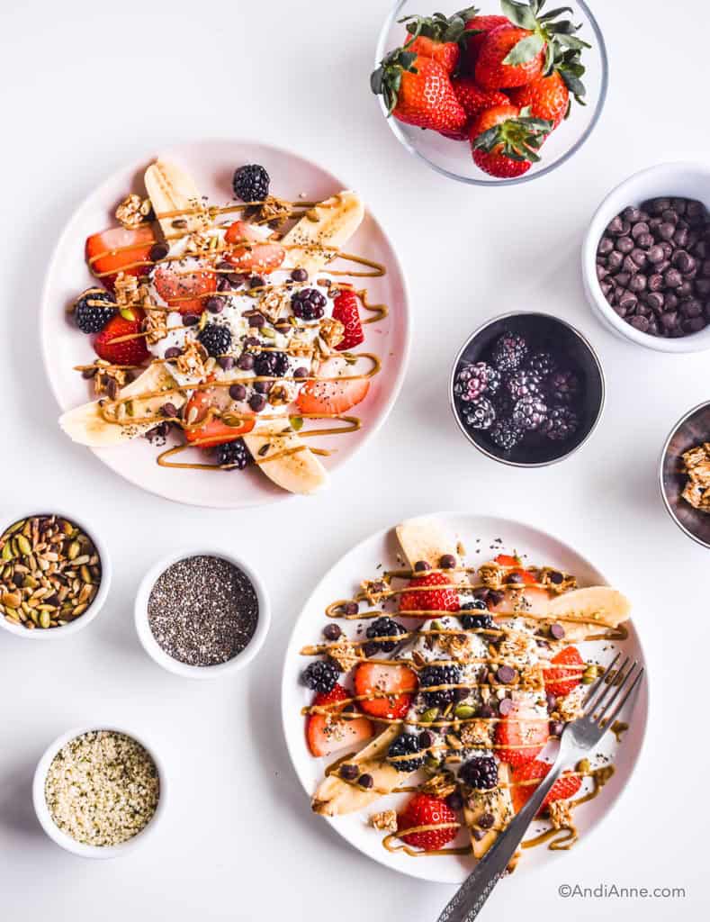 healthy banana splits on pink plates with a fork. small bowls of ingredients around the plates including white bols of pumpkin seeds, hemp seeds, chia seeds and chocolate chips.