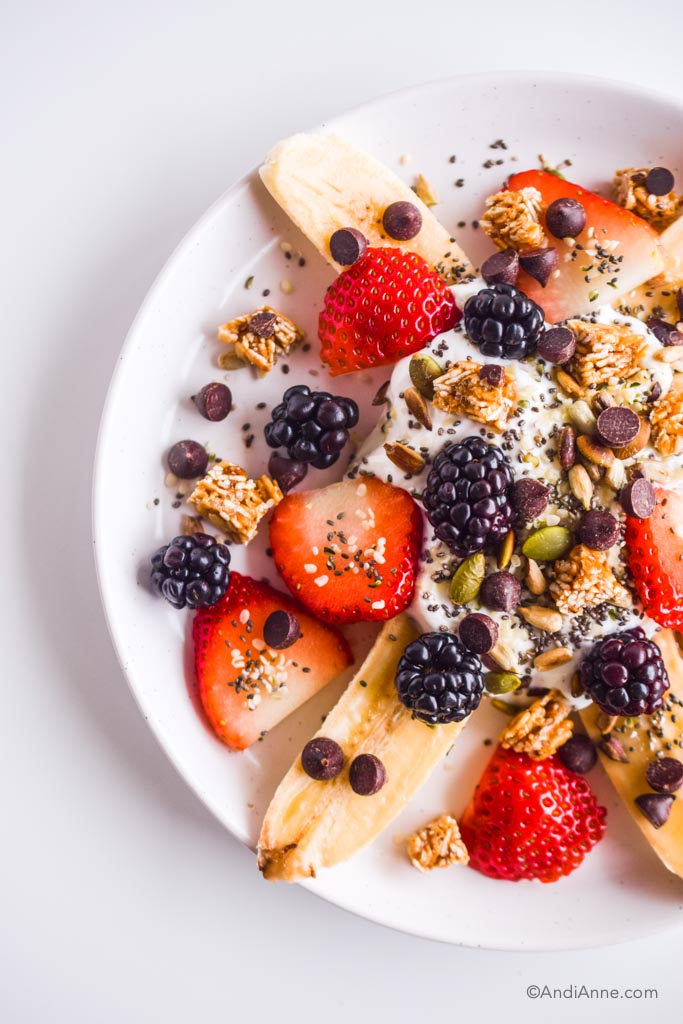 Detail of healthy banana split on white plate with strawberries, blackberries, seeds, and chocolate chips.