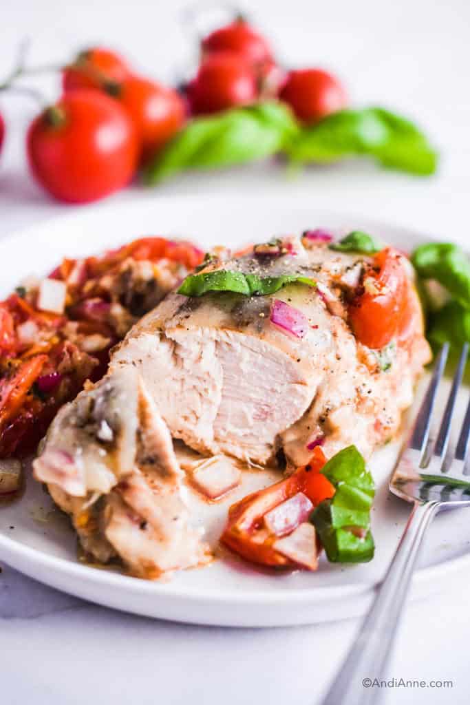 Chopped open chicken breast on plate with fork and tomatoes in background.