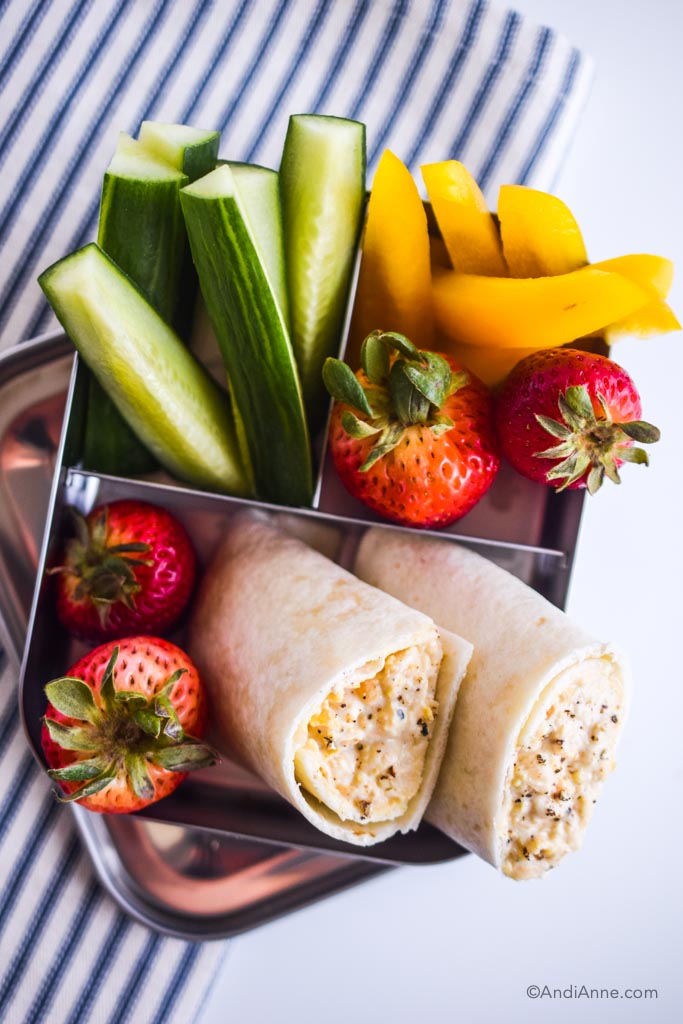 mashed chickpea wrap kids lunch in bento lunch box with sliced cucumber, sliced bell peppers, and strawberries. On a blue and white striped napkin.