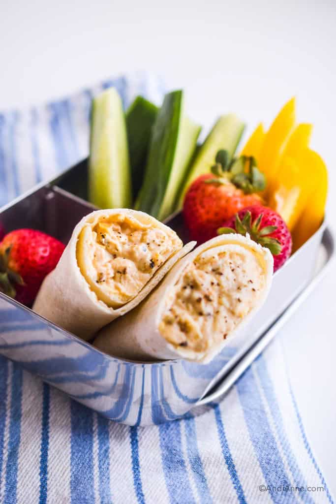 mashed chickpea wrap kids lunch in bento lunch box with sliced cucumber, sliced bell peppers, and strawberries. On a blue and white striped napkin.