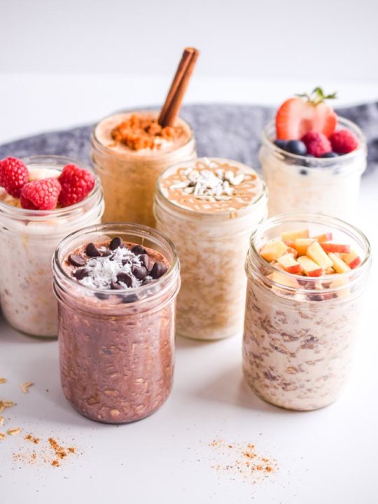 six overnight oats recipes in mason jars with fresh fruit toppings and dark napkin in background