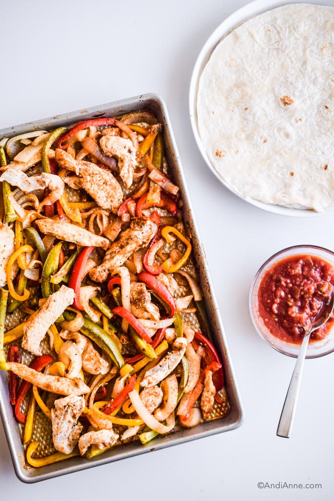sheet pan fajitas alongside a plate of flour tortilla wraps, and a glass bowl of salsa with a spoon in it.