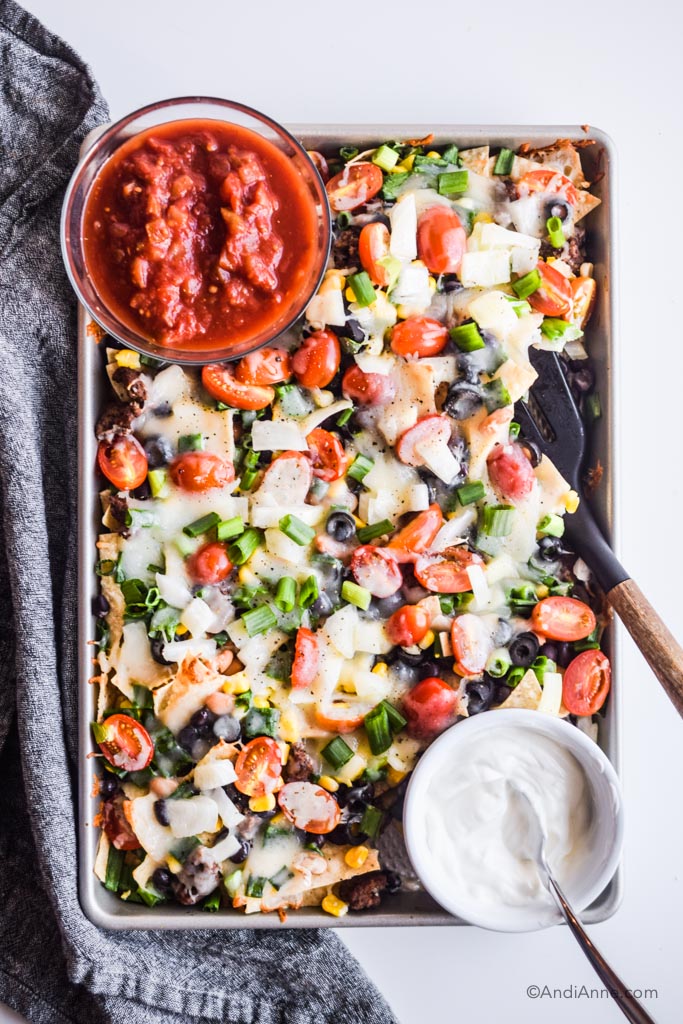 sheet pan nachos on baking sheet with bowl of salsa and bowl of sour cream. Spatula with wooden handle