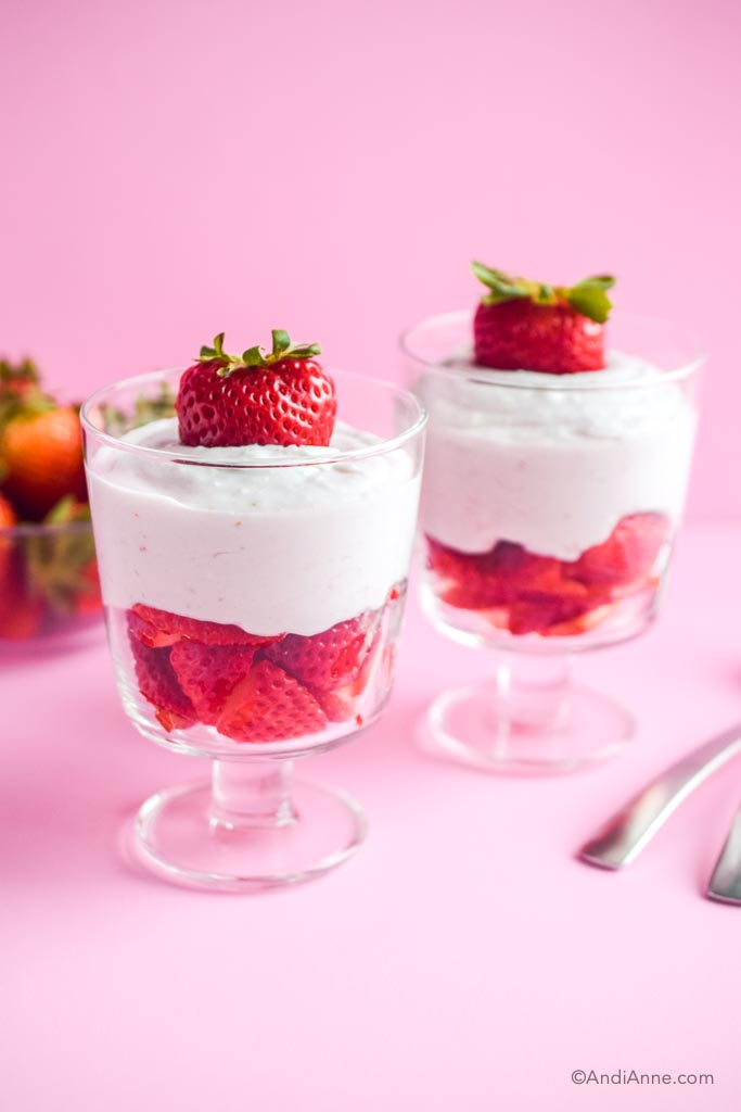 two strawberries and cream desserts in glasses with two spoons beside and a pink background
