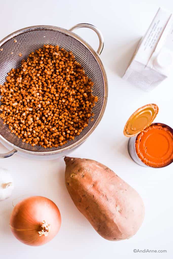 ingredients used for the soup including lentils in a metal strainer, a can of pumpkin puree, a large sweet potato, yellow onion, garlic, and carton of broth