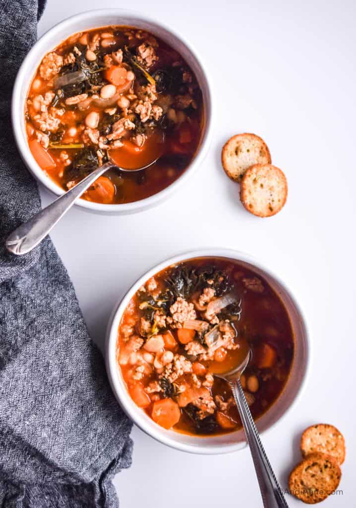 two white bowls of ground turkey soup with silver spoons. Two crackers and dark napkin beside it.